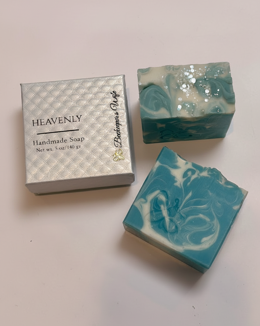 Two handmade soap bars with teal, white, and glitter swirls displayed next to a silver gift box labeled HEAVENLY and Butterscotch Pie on a light background.