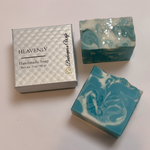 Two handmade soap bars with teal, white, and glitter swirls displayed next to a silver gift box labeled HEAVENLY and Butterscotch Pie on a light background.