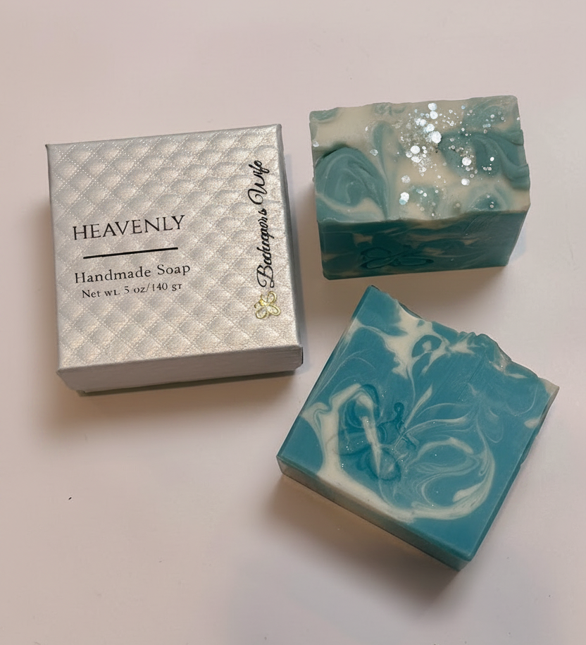 Two handmade soap bars with teal, white, and glitter swirls displayed next to a silver gift box labeled HEAVENLY and Butterscotch Pie on a light background.
