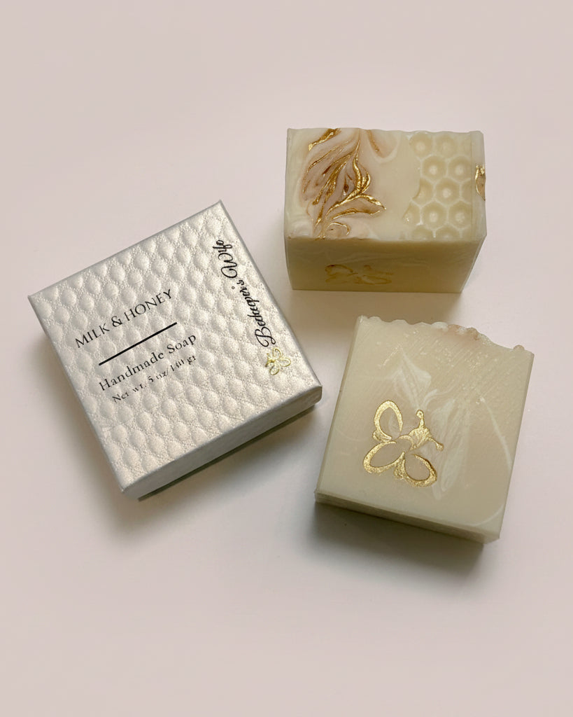 Two handmade soap bars with gold foil accents and a silver textured gift box labeled 'Milk & Honey Handmade Soap' on a light surface.