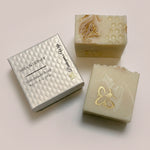 Two handmade soap bars with gold foil accents and a silver textured gift box labeled 'Milk & Honey Handmade Soap' on a light surface.