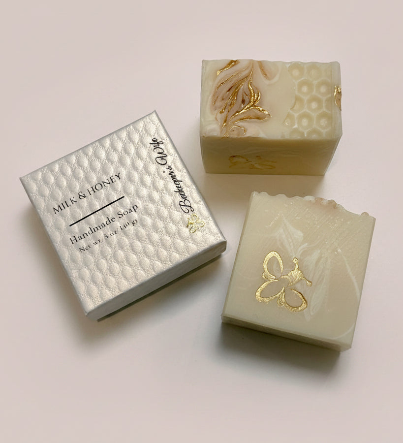 Two handmade soap bars with gold foil accents and a silver textured gift box labeled 'Milk & Honey Handmade Soap' on a light surface.