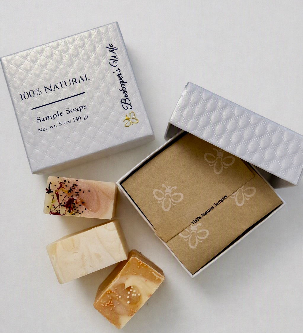 Three artisanal bar soaps displayed next to their packaging on a light gray background. The soaps feature natural, earthy tones with some speckled details.