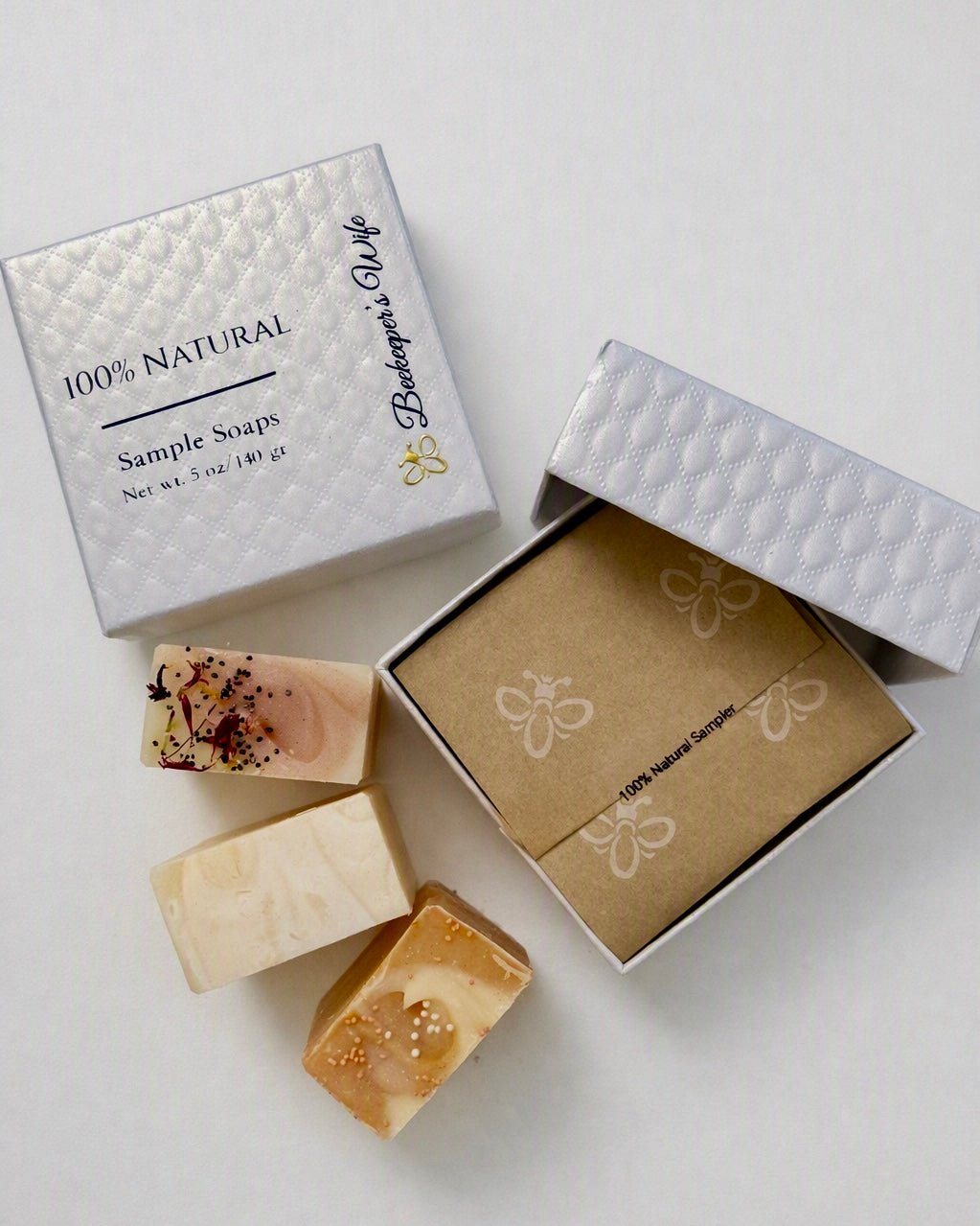 Three artisanal bar soaps displayed next to their packaging on a light gray background. The soaps feature natural, earthy tones with some speckled details.