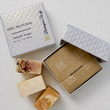 Three artisanal bar soaps displayed next to their packaging on a light gray background. The soaps feature natural, earthy tones with some speckled details.