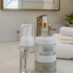 A skincare set featuring a clear bottle with a white pump dispenser, a stack of two frosted jars labeled 'HONEY + ALOE HYDRATION CREAM' and 'HONEY + ALOE JELLY MASK', and a small dropper bottle with a gold collar, all on a white surface with a decorative gray background.