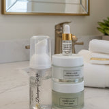 A skincare set featuring a clear bottle with a white pump dispenser, a stack of two frosted jars labeled 'HONEY + ALOE HYDRATION CREAM' and 'HONEY + ALOE JELLY MASK', and a small dropper bottle with a gold collar, all on a white surface with a decorative gray background.