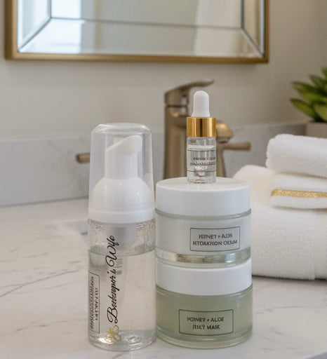 A skincare set featuring a clear bottle with a white pump dispenser, a stack of two frosted jars labeled 'HONEY + ALOE HYDRATION CREAM' and 'HONEY + ALOE JELLY MASK', and a small dropper bottle with a gold collar, all on a white surface with a decorative gray background.