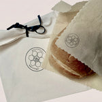 A close-up of a round bread roll wrapped in a translucent parchment sleeve with a circular logo, placed beside a white fabric dust bag tied with a black ribbon. The bread and packaging display the brand name 'BEEUSABLE'.