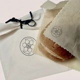 A close-up of a round bread roll wrapped in a translucent parchment sleeve with a circular logo, placed beside a white fabric dust bag tied with a black ribbon. The bread and packaging display the brand name 'BEEUSABLE'.