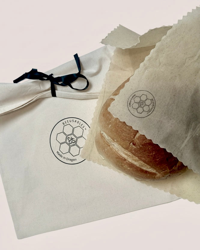 A close-up of a round bread roll wrapped in a translucent parchment sleeve with a circular logo, placed beside a white fabric dust bag tied with a black ribbon. The bread and packaging display the brand name 'BEEUSABLE'.