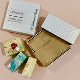 Three decorative handmade soap bars arranged beside two gift boxes on a soft blush background. One closed box is labeled 'DESIGNER Sample Soaps' with 'Beekeeper's Wife' printed vertically, and the open box shows kraft paper lining.