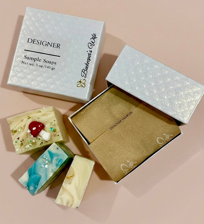 Three decorative handmade soap bars arranged beside two gift boxes on a soft blush background. One closed box is labeled 'DESIGNER Sample Soaps' with 'Beekeeper's Wife' printed vertically, and the open box shows kraft paper lining.