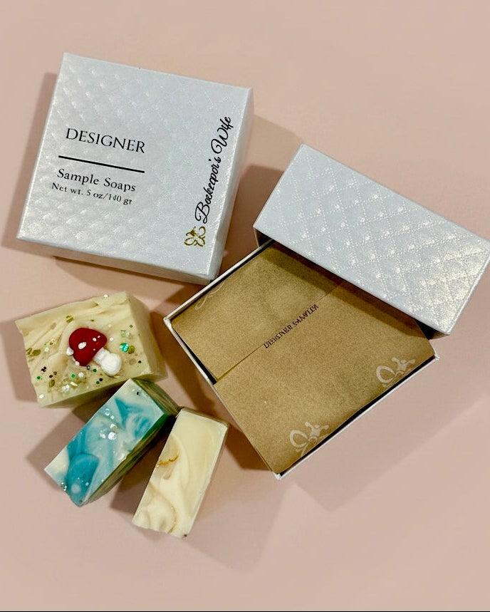 Three decorative handmade soap bars arranged beside two gift boxes on a soft blush background. One closed box is labeled 'DESIGNER Sample Soaps' with 'Beekeeper's Wife' printed vertically, and the open box shows kraft paper lining.