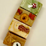 Four rectangular artisanal soaps stacked vertically, each decorated with autumn-themed motifs including a mushroom, a pumpkin, honeycomb, and maple leaves, on a plain light background.