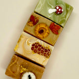 Four rectangular artisanal soaps stacked vertically, each decorated with autumn-themed motifs including a mushroom, a pumpkin, honeycomb, and maple leaves, on a plain light background.