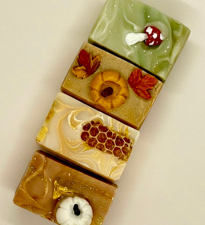 Four rectangular artisanal soaps stacked vertically, each decorated with autumn-themed motifs including a mushroom, a pumpkin, honeycomb, and maple leaves, on a plain light background.