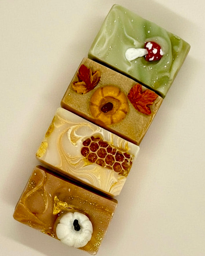 Four rectangular artisanal soaps stacked vertically, each decorated with autumn-themed motifs including a mushroom, a pumpkin, honeycomb, and maple leaves, on a plain light background.