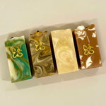 Four rectangular marbled soap bars arranged in a row on a light beige background. Each soap has a different color combination: green and white, tan and brown, cream and beige, and brown and white, with a small gold butterfly design on the front of each bar.