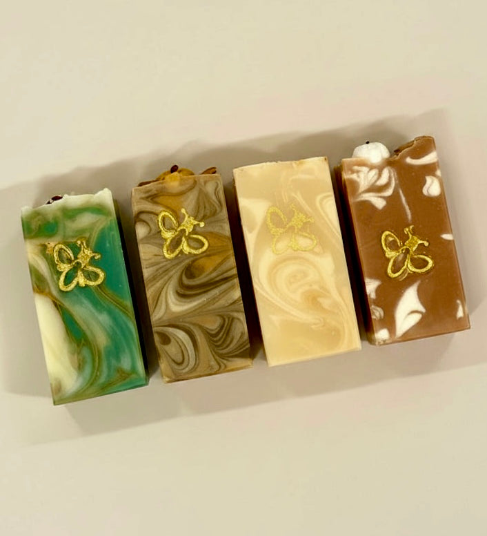 Four rectangular marbled soap bars arranged in a row on a light beige background. Each soap has a different color combination: green and white, tan and brown, cream and beige, and brown and white, with a small gold butterfly design on the front of each bar.