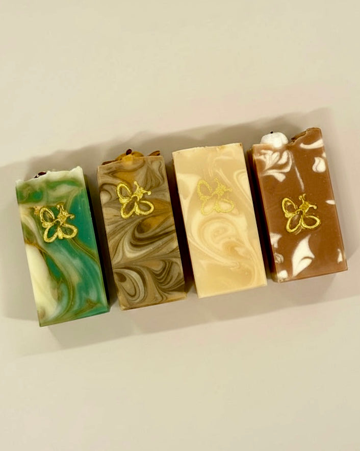 Four rectangular marbled soap bars arranged in a row on a light beige background. Each soap has a different color combination: green and white, tan and brown, cream and beige, and brown and white, with a small gold butterfly design on the front of each bar.