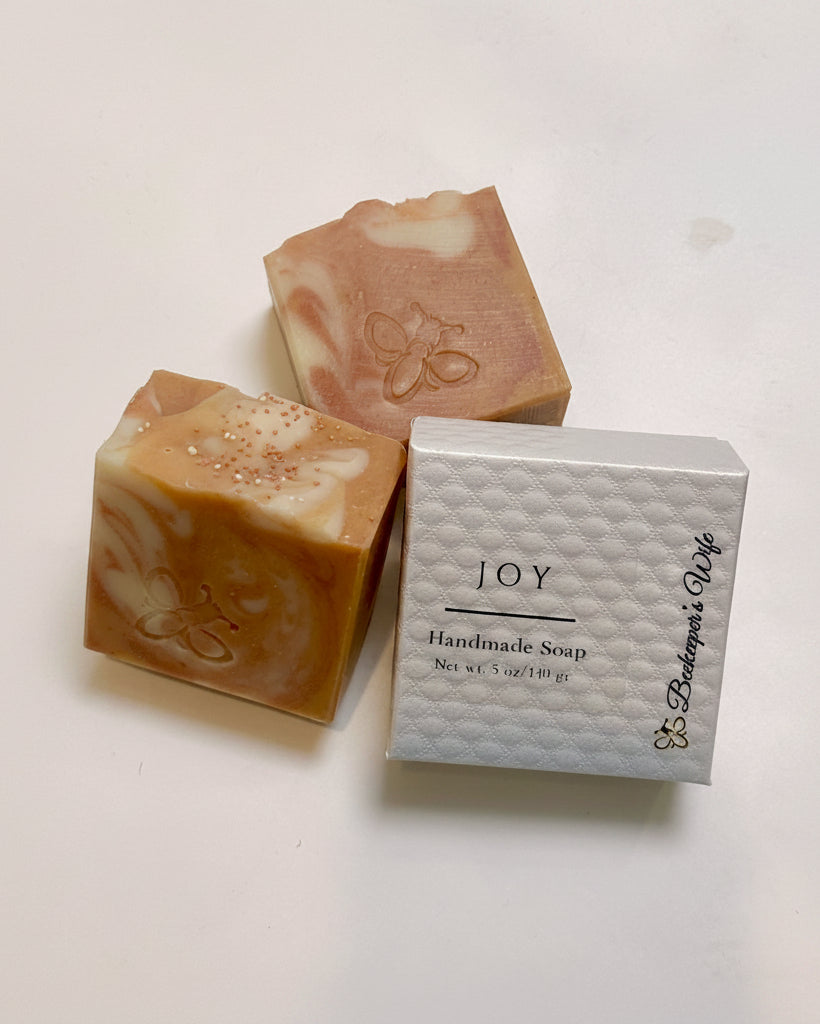 Three handmade soap bars with peach, cream, and pink marbled swirls are displayed next to their white textured packaging box labeled 'JOY Handmade Soap' by 'Bakerbee's Wife' on a white surface.