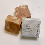 Three handmade soap bars with peach, cream, and pink marbled swirls are displayed next to their white textured packaging box labeled 'JOY Handmade Soap' by 'Bakerbee's Wife' on a white surface.