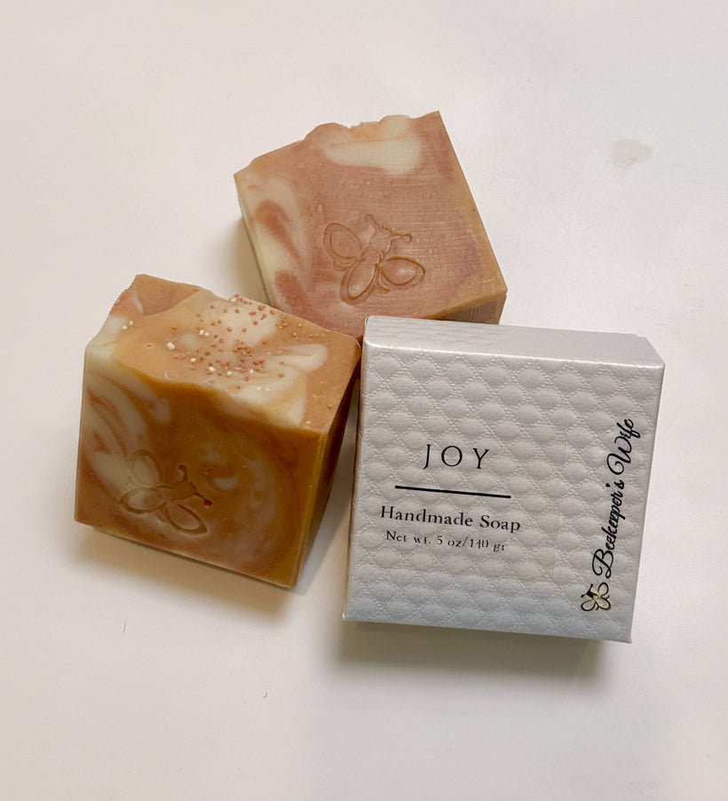 Three handmade soap bars with peach, cream, and pink marbled swirls are displayed next to their white textured packaging box labeled 'JOY Handmade Soap' by 'Bakerbee's Wife' on a white surface.