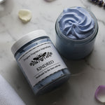 Jar of blue whipped body butter on it's side next to an open jar showing the contents.