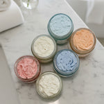Six small jars with colorful whipped body butter on a white surface