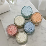 Six small jars with colorful whipped body butter on a white surface