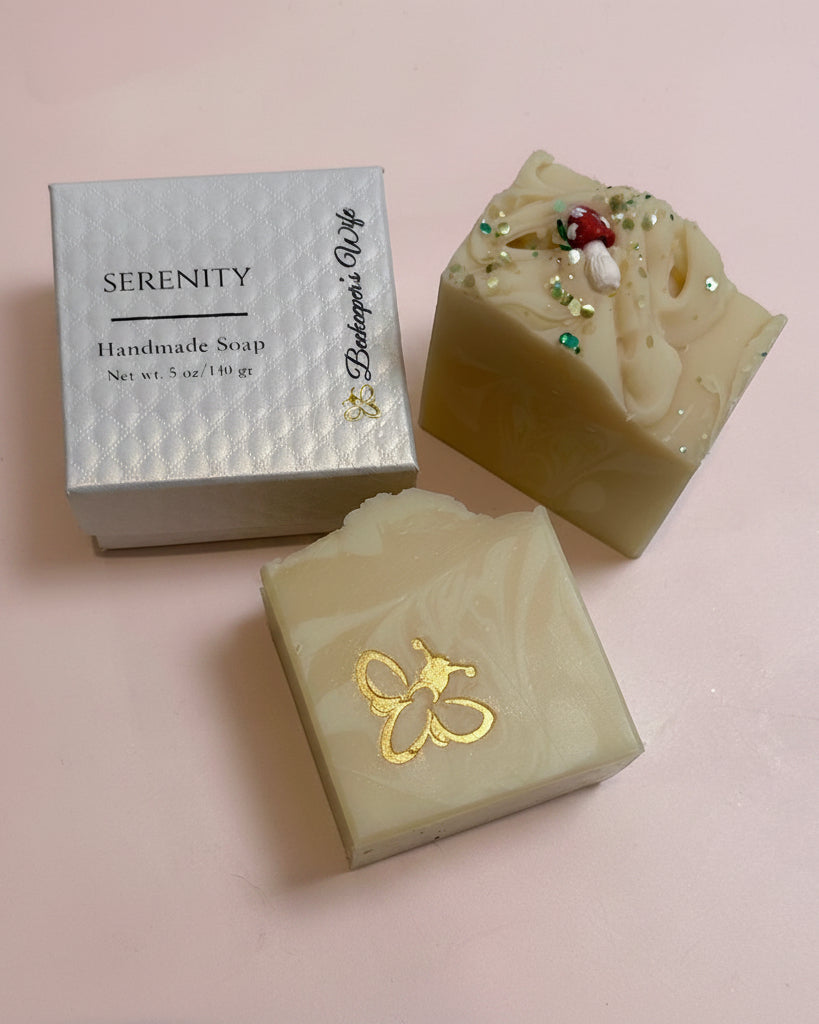 Two handmade soap bars and their packaging box on a white surface. One bar is a light cream color with a gold foil bee design, and the other is a marbled beige and yellow soap topped with green and red glitter.