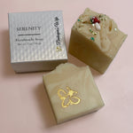 Two handmade soap bars and their packaging box on a white surface. One bar is a light cream color with a gold foil bee design, and the other is a marbled beige and yellow soap topped with green and red glitter.