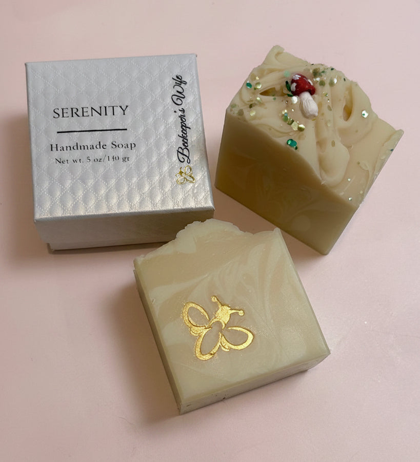 Two handmade soap bars and their packaging box on a white surface. One bar is a light cream color with a gold foil bee design, and the other is a marbled beige and yellow soap topped with green and red glitter.