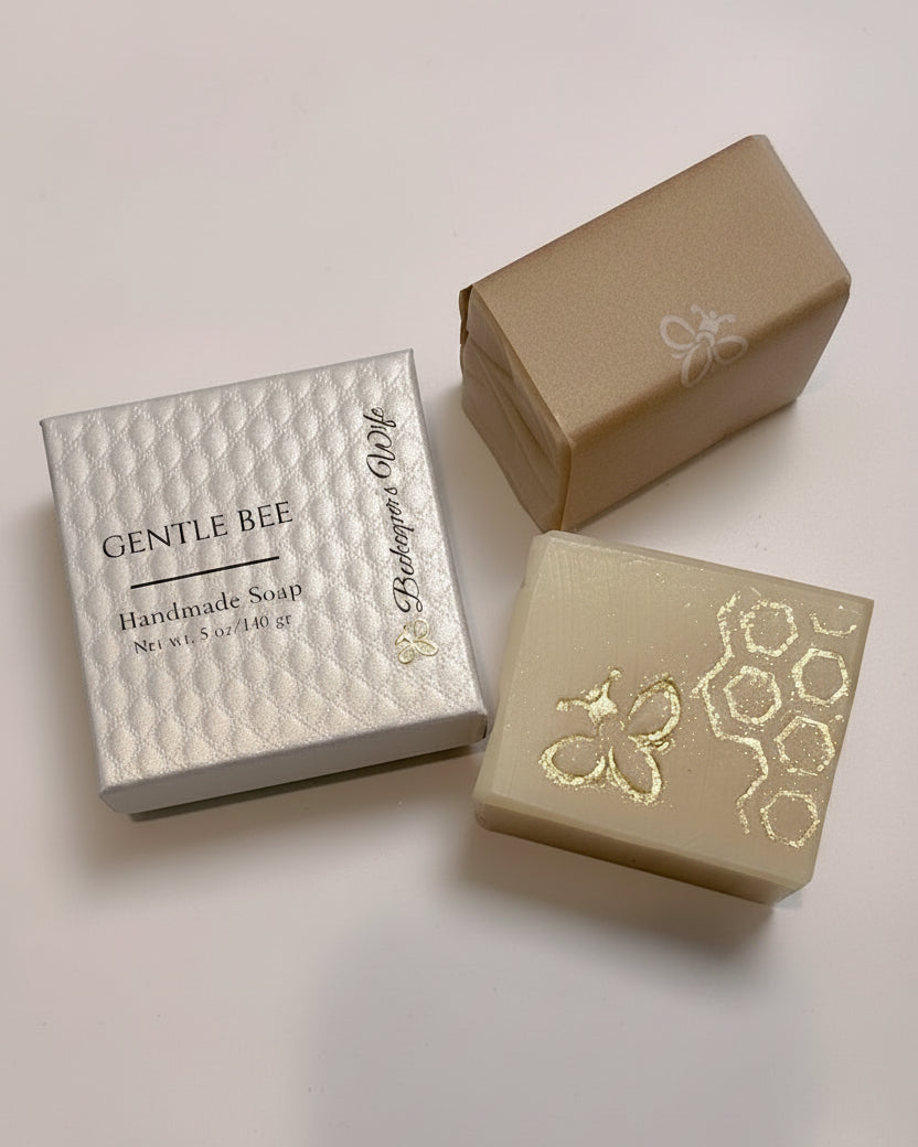 Two rectangular bars of handmade soap displayed with their packaging on a light off-white background. One soap bar is a warm cream color with gold foil bee and honeycomb patterns, paired with a textured white box labeled 'GENTLE BEE Handmade Soap'.