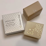 Two rectangular bars of handmade soap displayed with their packaging on a light off-white background. One soap bar is a warm cream color with gold foil bee and honeycomb patterns, paired with a textured white box labeled 'GENTLE BEE Handmade Soap'.