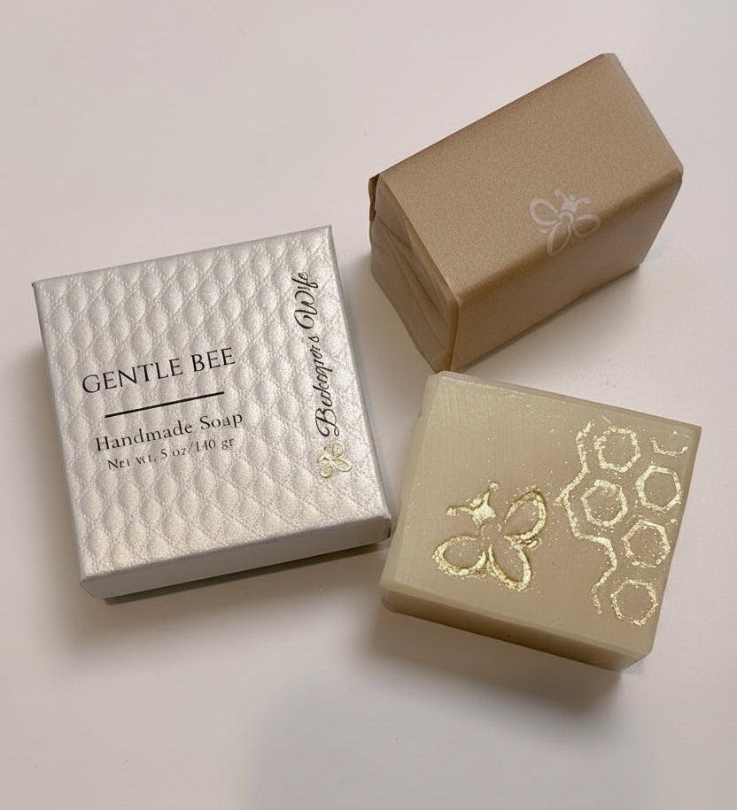 Two rectangular bars of handmade soap displayed with their packaging on a light off-white background. One soap bar is a warm cream color with gold foil bee and honeycomb patterns, paired with a textured white box labeled 'GENTLE BEE Handmade Soap'.