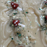 A close-up of a textured cream-colored soap bar decorated with small red and white mushroom shapes and scattered green and silver glitter.