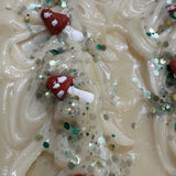 A close-up of a textured cream-colored soap bar decorated with small red and white mushroom shapes and scattered green and silver glitter.