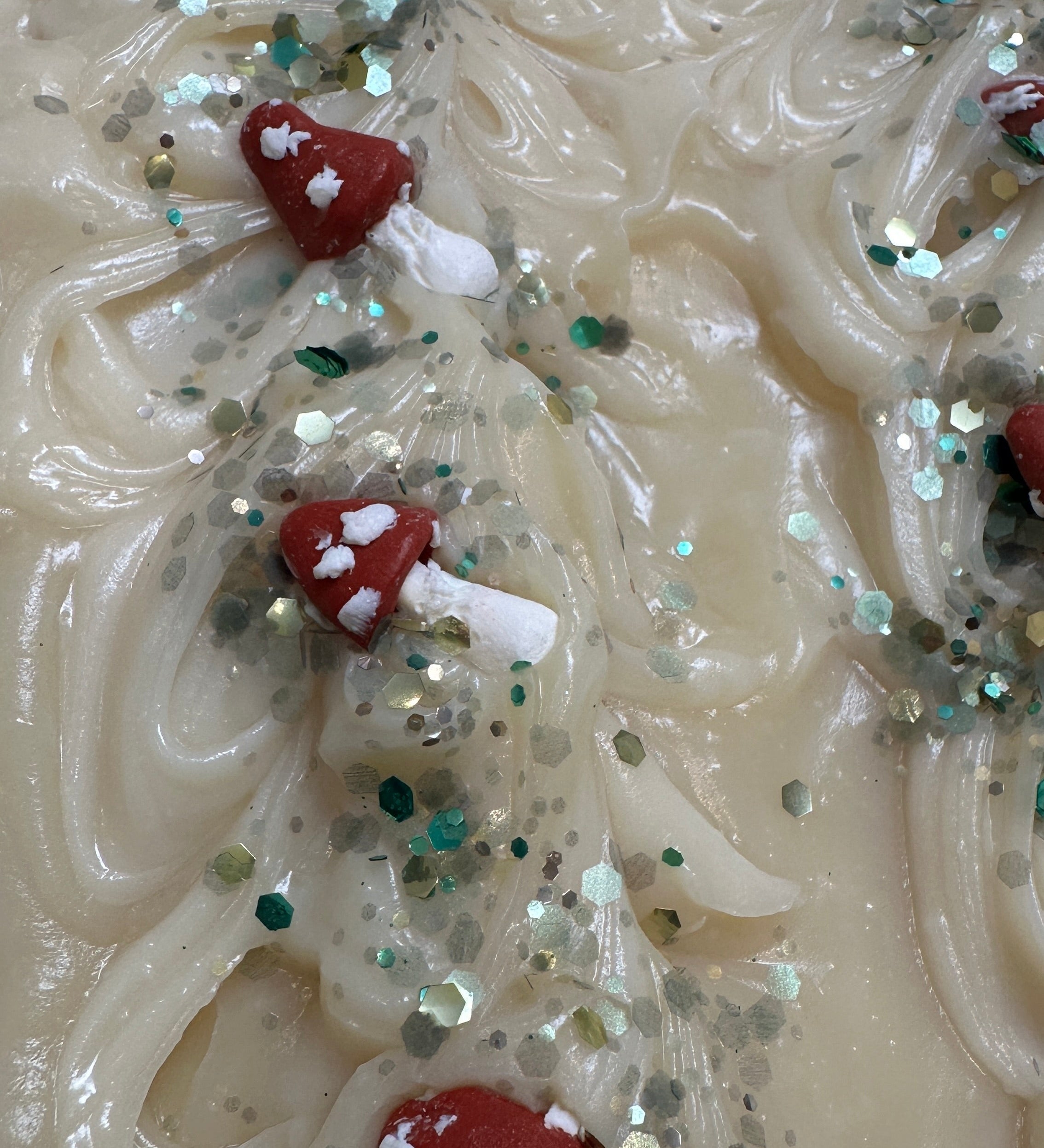 A close-up of a textured cream-colored soap bar decorated with small red and white mushroom shapes and scattered green and silver glitter.