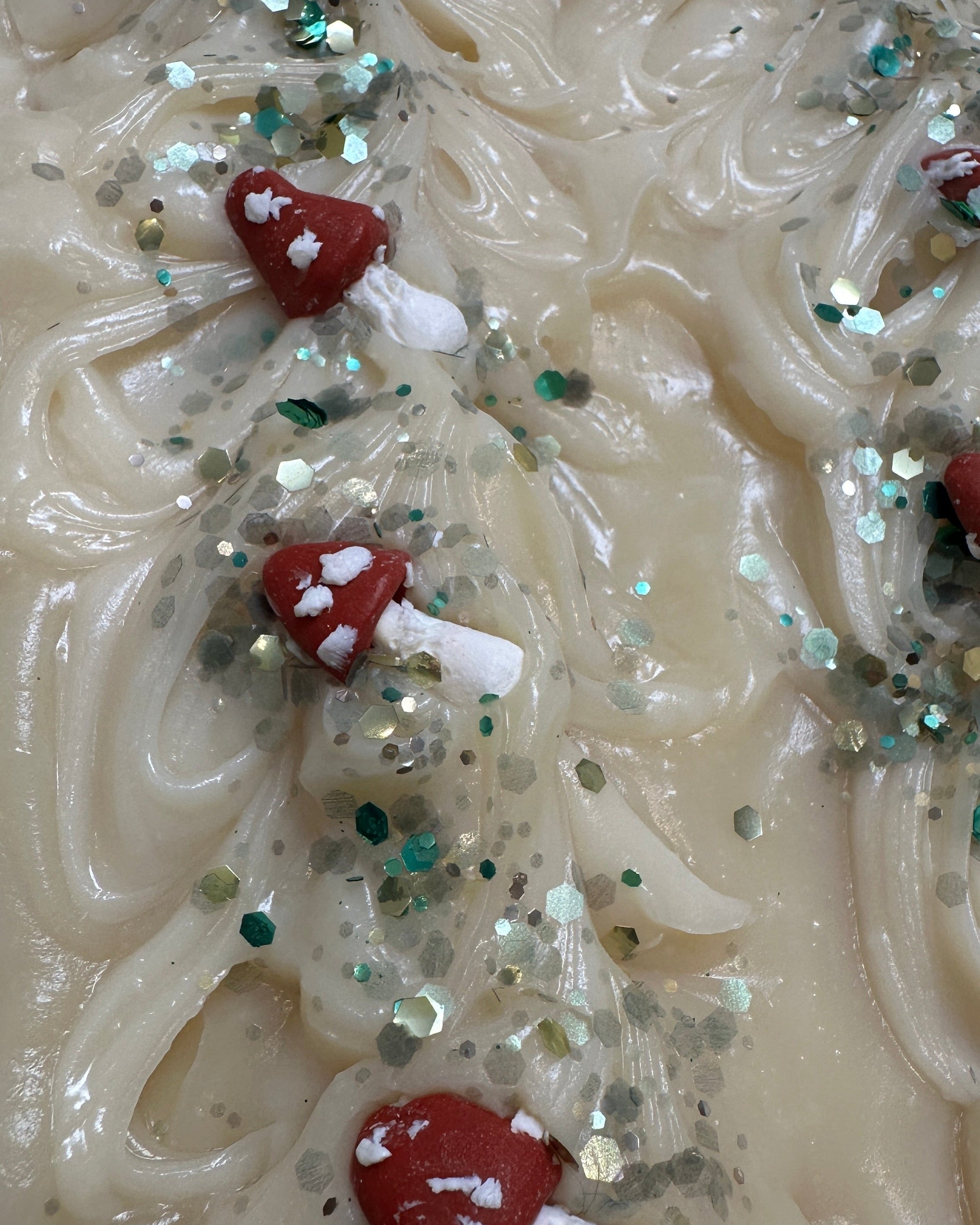 A close-up of a textured cream-colored soap bar decorated with small red and white mushroom shapes and scattered green and silver glitter.